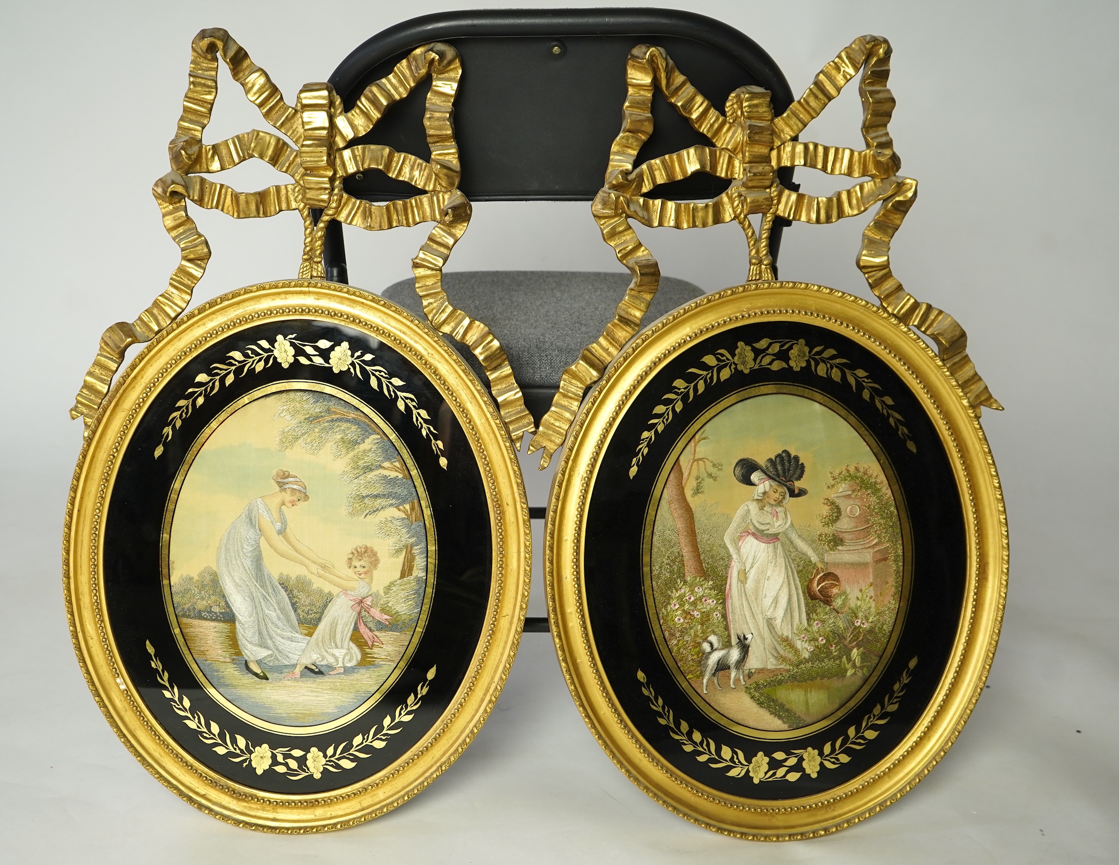 A pair of early 20th century oval silk embroideries, one of a mother and child dancing in a garden, the other of a lady in a large brimmed hat, watering flowers in by a classical stone urn, watched by her dog. Framed in